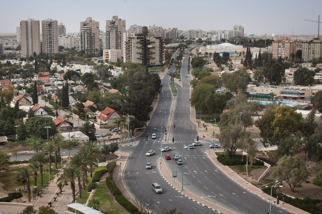 Aerial view of Be'er-Sheva showing modern high-rise apartment buildings, palm trees, and a wide main road with traffic.
