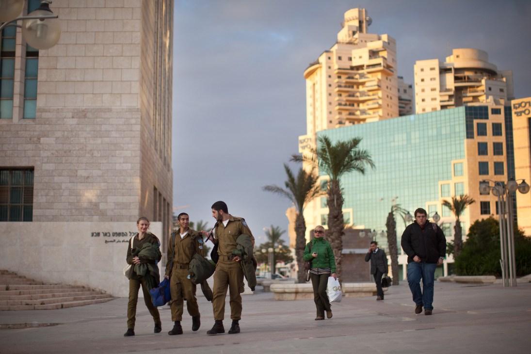 Israeli soldiers in uniform walking through Be'er-Sheva city center with palm trees and modern buildings in background.