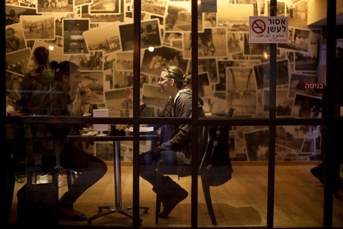 Two women sit at a table inside Ringelblum café, with historical photographs covering the walls behind them.