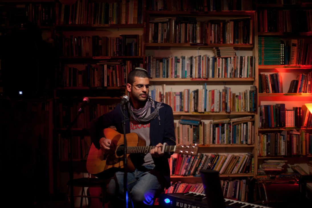 Ohad Darshan performs with an acoustic guitar on stage at Ashan Hazman bar in Be'er-Sheva, with illuminated bookshelves behind him.