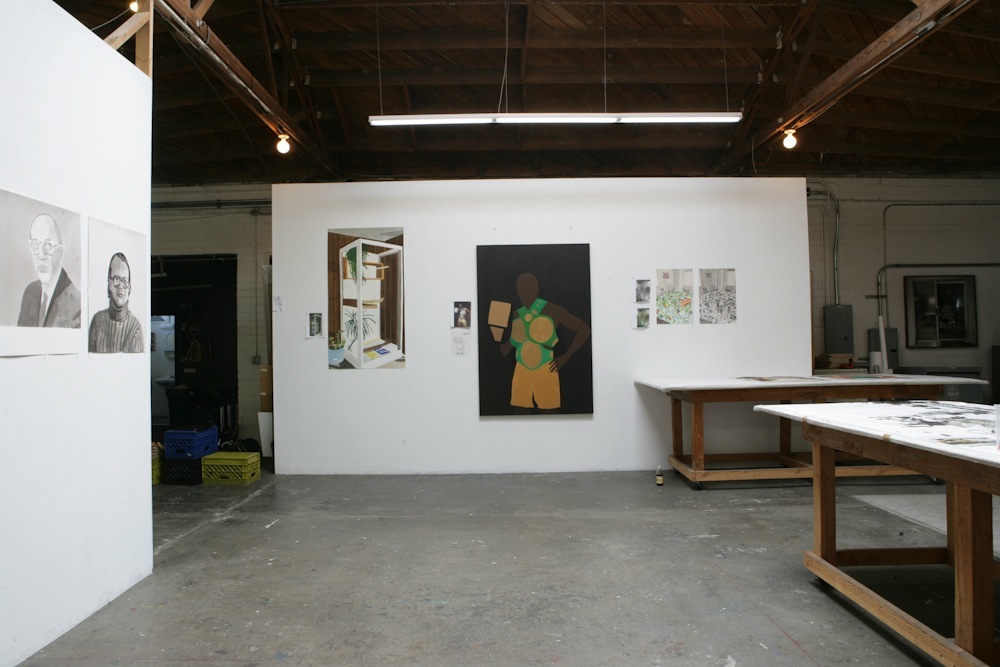 Interior of an artist's studio with white walls displaying various artworks and wooden work tables.