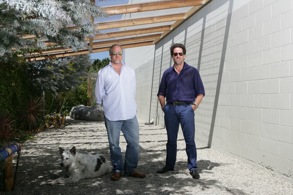 Two men stand in a sunlit courtyard with white walls and wooden beams; a dog rests in the foreground.