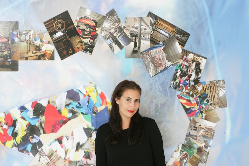 Lauri Firstenberg, director of LAXART, poses in front of a light blue wall with photographs and colorful collage artwork arranged in a circular pattern.