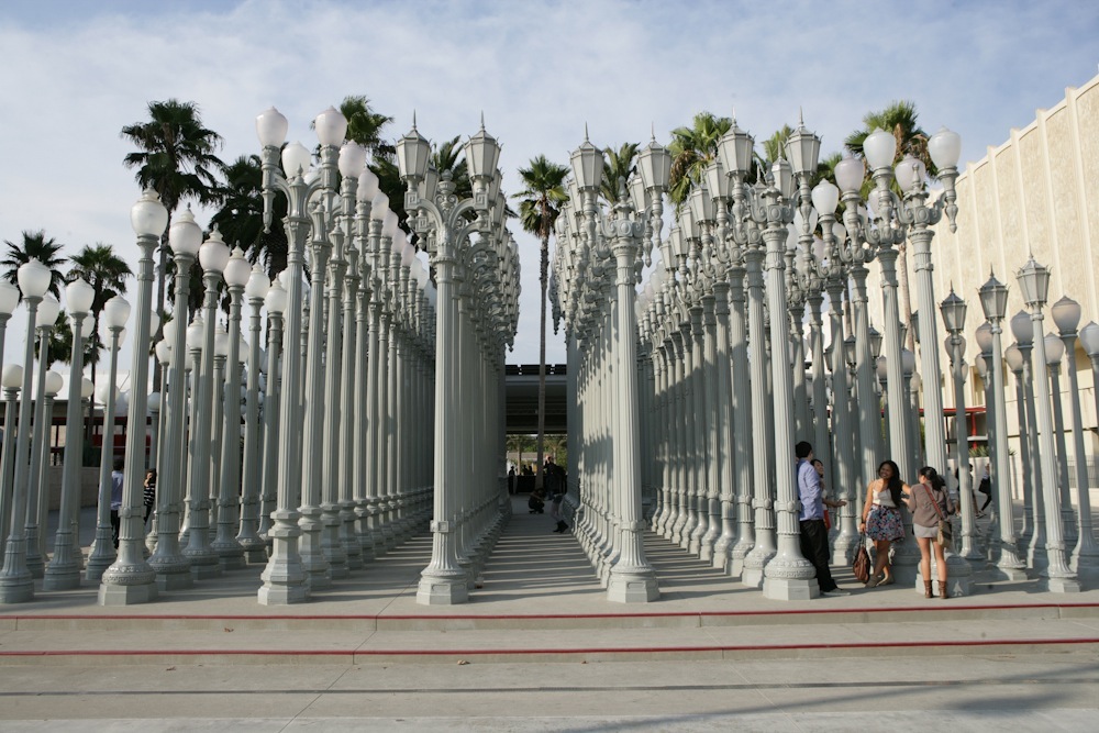 Chris Burden's Urban Light installation featuring rows of white ornamental street lamps at LACMA with palm trees and visitors.