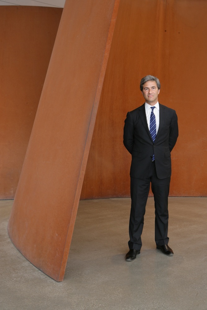 Michael Govan, director of LACMA, stands in a dark suit before a large rust-colored curved sculpture.