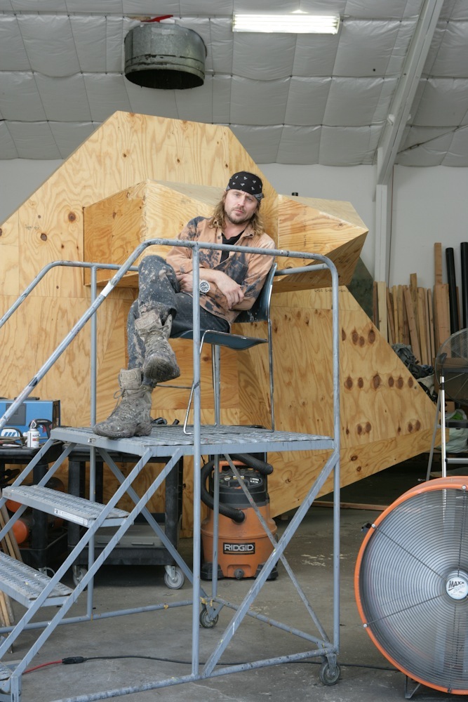 Artist Sterling Ruby sits on a metal scaffold in front of a large wooden sculptural form in his industrial studio.