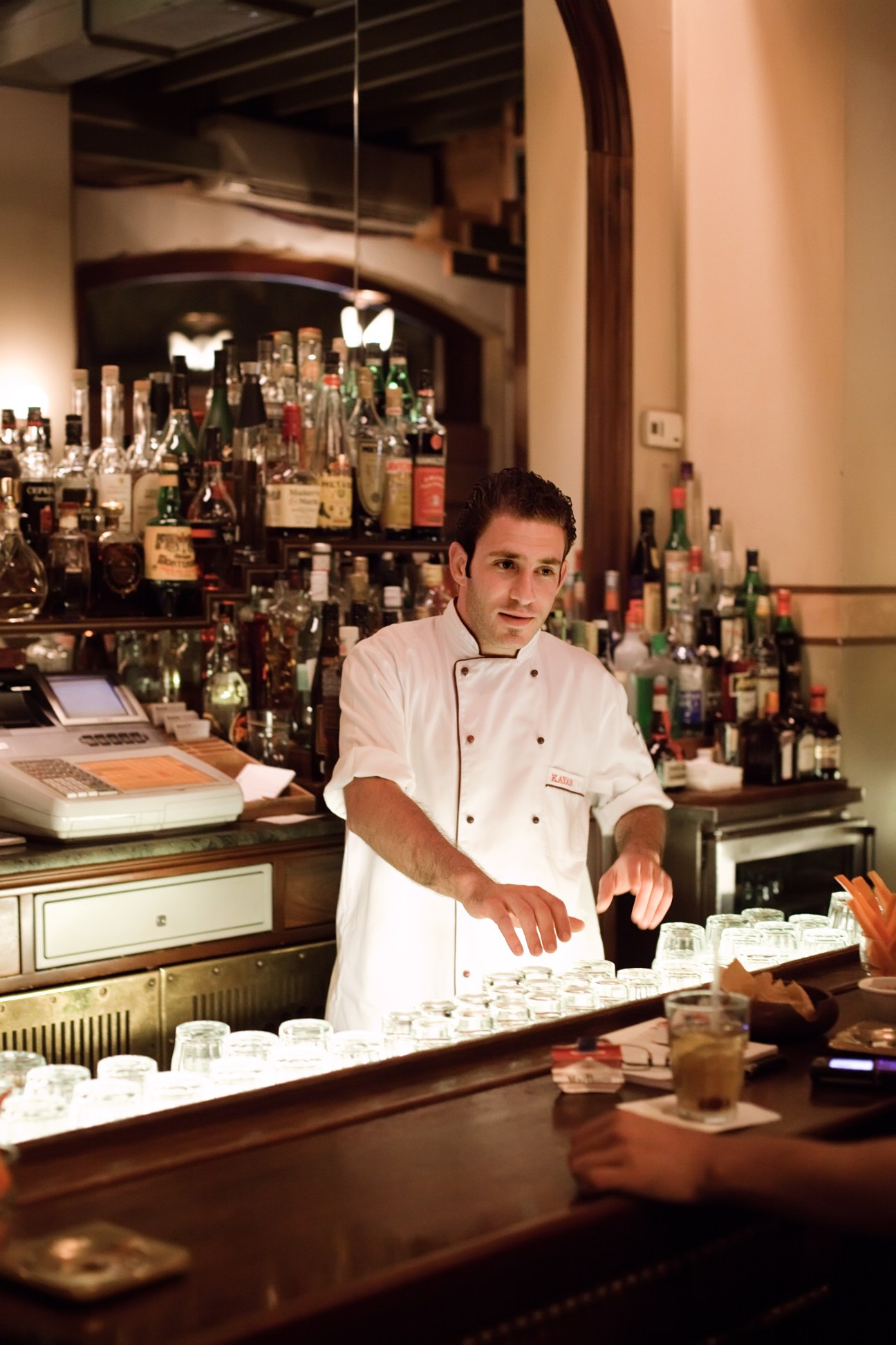 Barman in white chef's coat working behind bar with bottles of spirits displayed in background