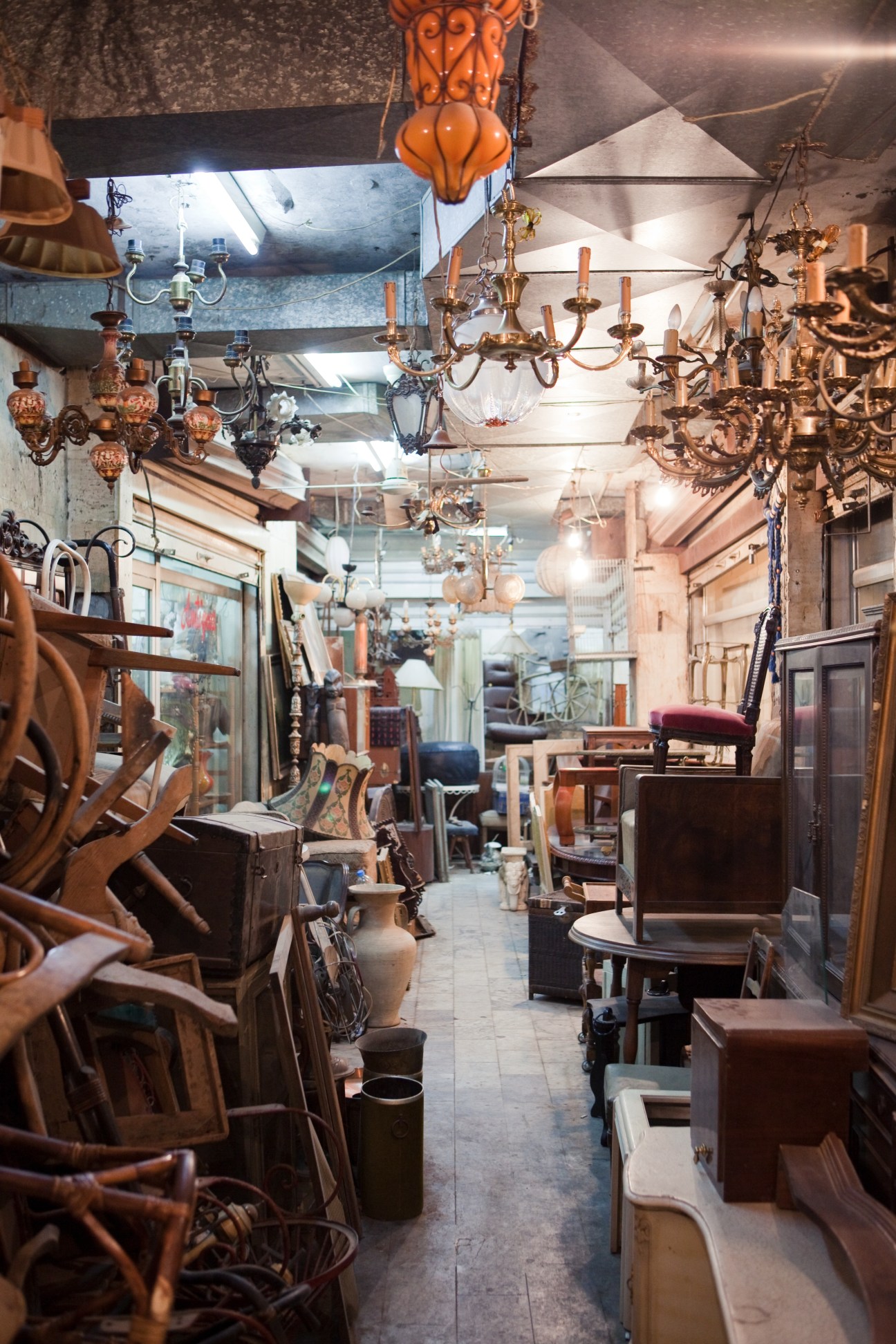 Interior of an antique shop displaying vintage chandeliers, furniture and decorative items hanging from ceiling and arranged throughout narrow space.