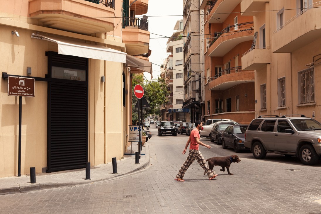 A person walks a brown dog down a quiet Beirut street lined with terracotta-colored residential buildings.