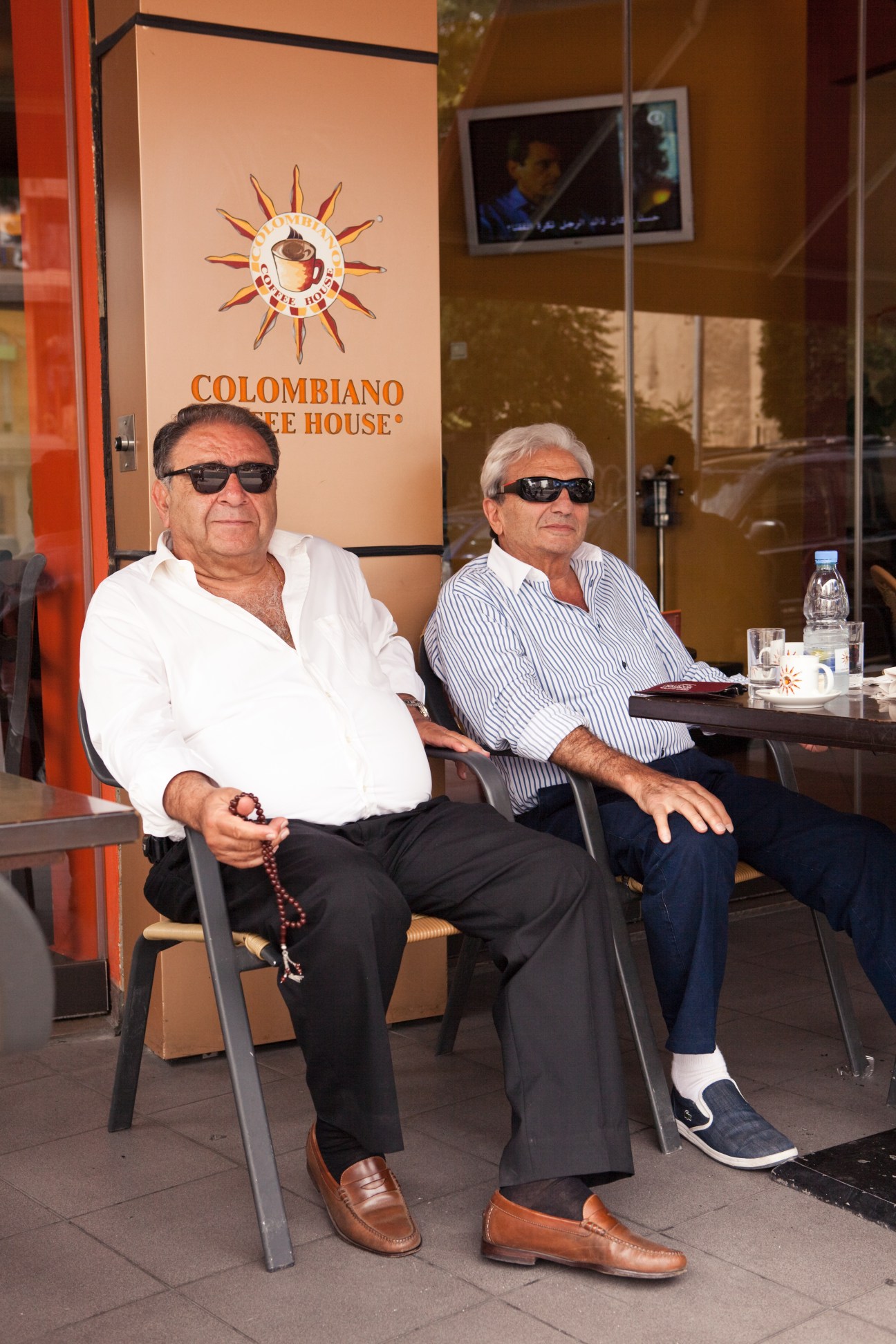 Two elderly men wearing sunglasses sit outside Colombiano Coffee House with a coffee cup and sun logo visible on the storefront behind them.