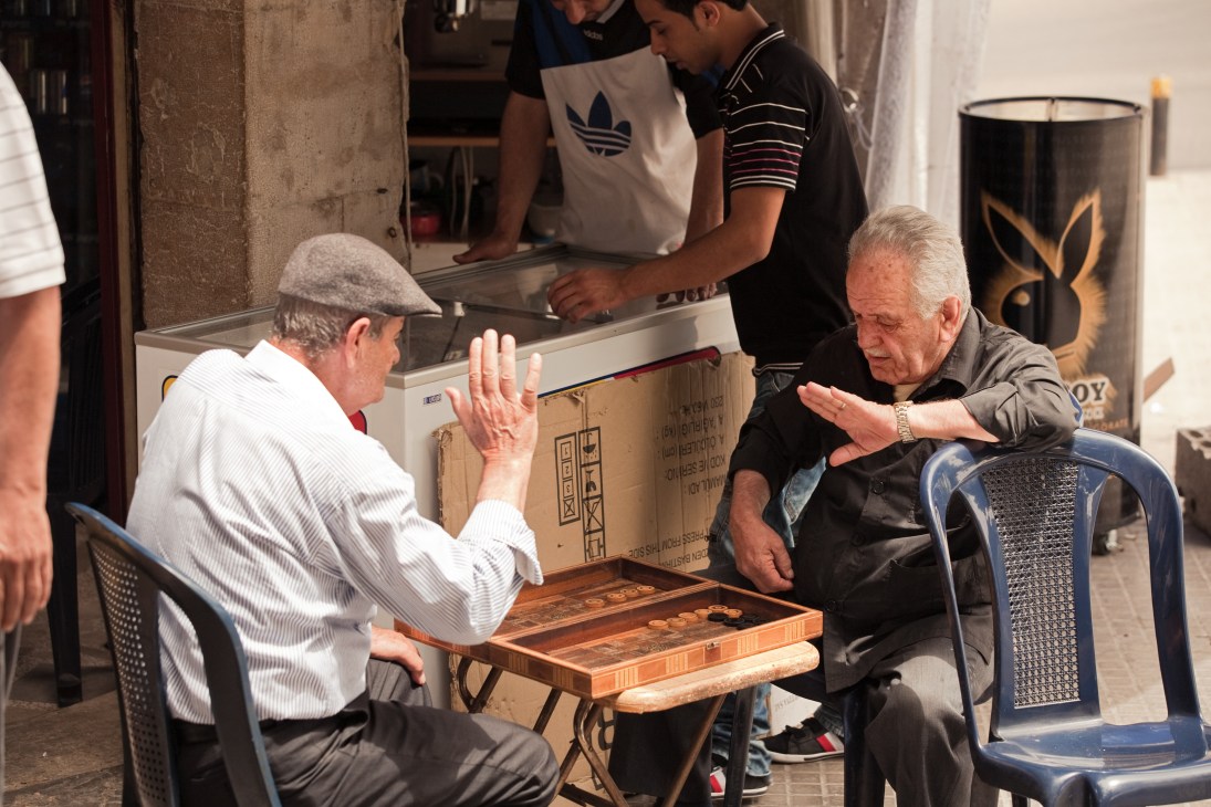 Two men play backgammon on a street in Ashrafieh, Beirut, with a shop and pedestrians visible in the background.