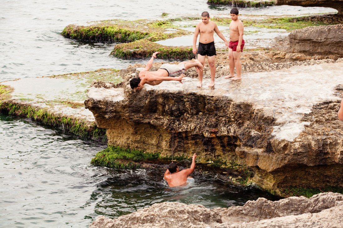 Young men diving and swimming off rocky outcrops at Beirut's Corniche waterfront.