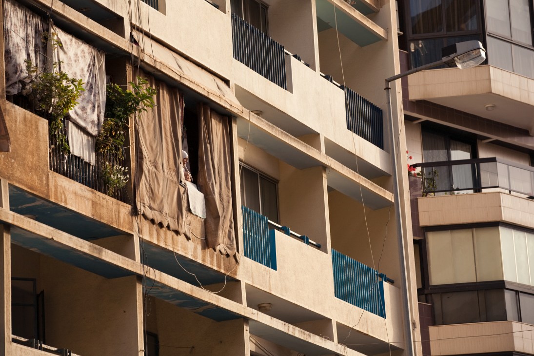 Weathered residential building facade in Manara with beige concrete, blue window grilles, and laundry hanging from balconies.