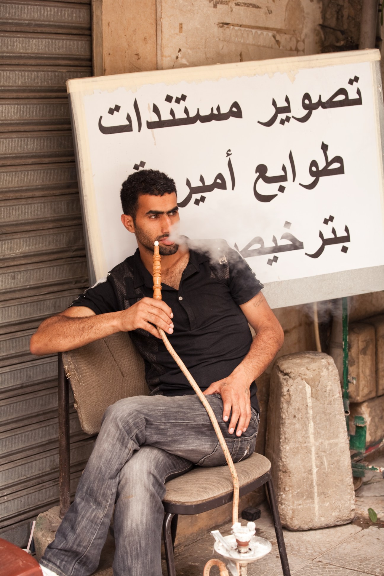 A man sits on a chair smoking a pipe in front of an Arabic signage in Ashrafieh, Beirut.