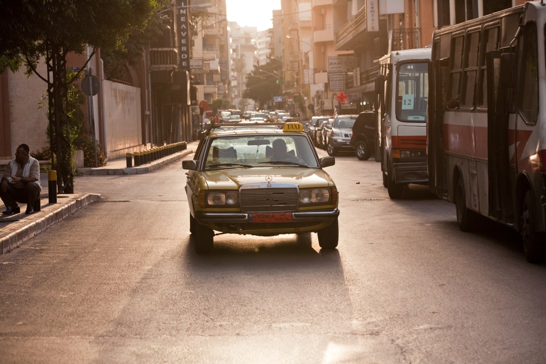A yellow Mercedes taxi drives down a sunny Beirut street lined with buildings and a bus.