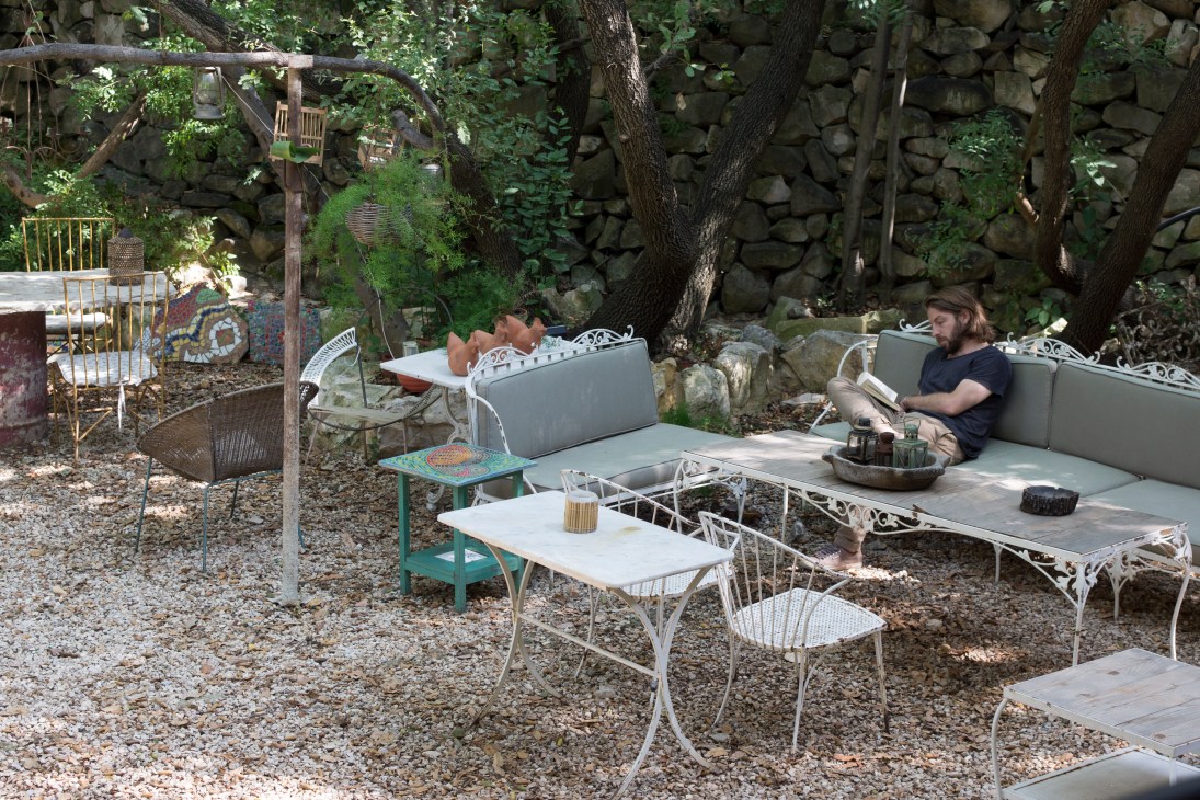 A guest relaxes on a gray sofa in Beit al Batroun's patio surrounded by vintage furniture and stone walls.