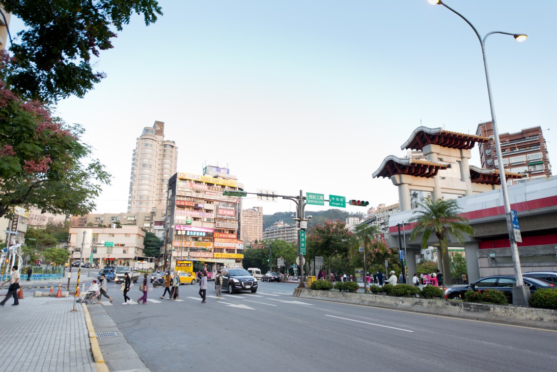Busy street scene in Beitou with mixed-height buildings, shops, vehicles, and pedestrians crossing.