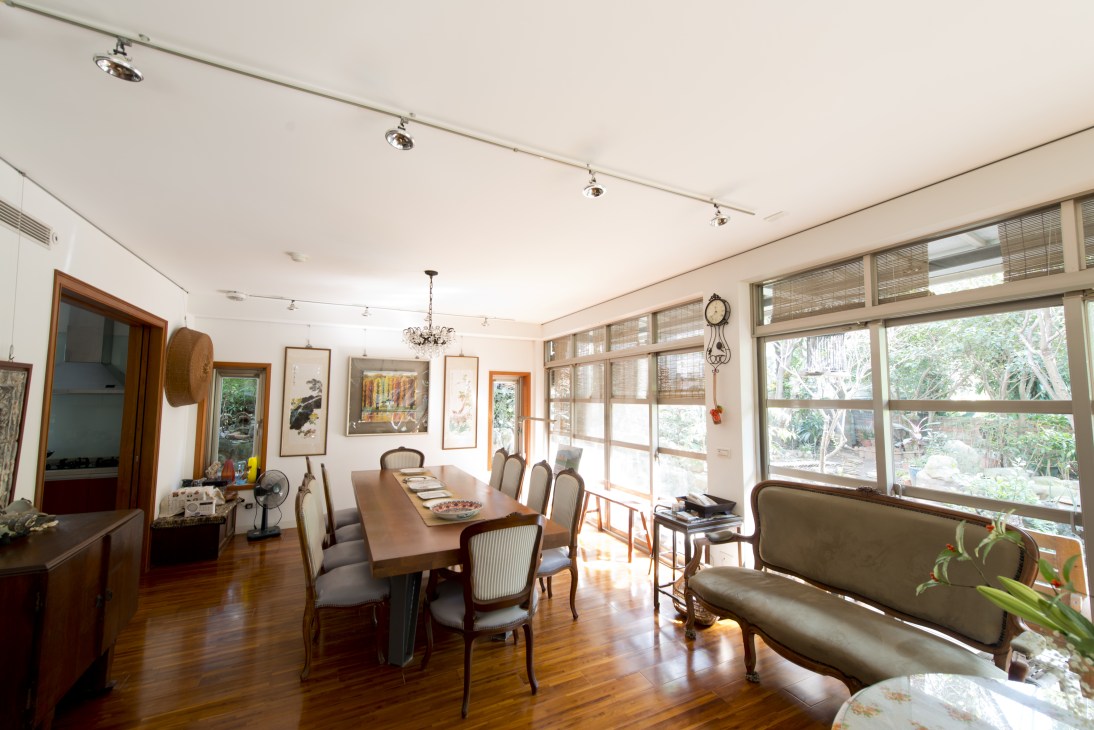 Bright living room with wooden dining table, framed artwork on walls, and large windows overlooking greenery.