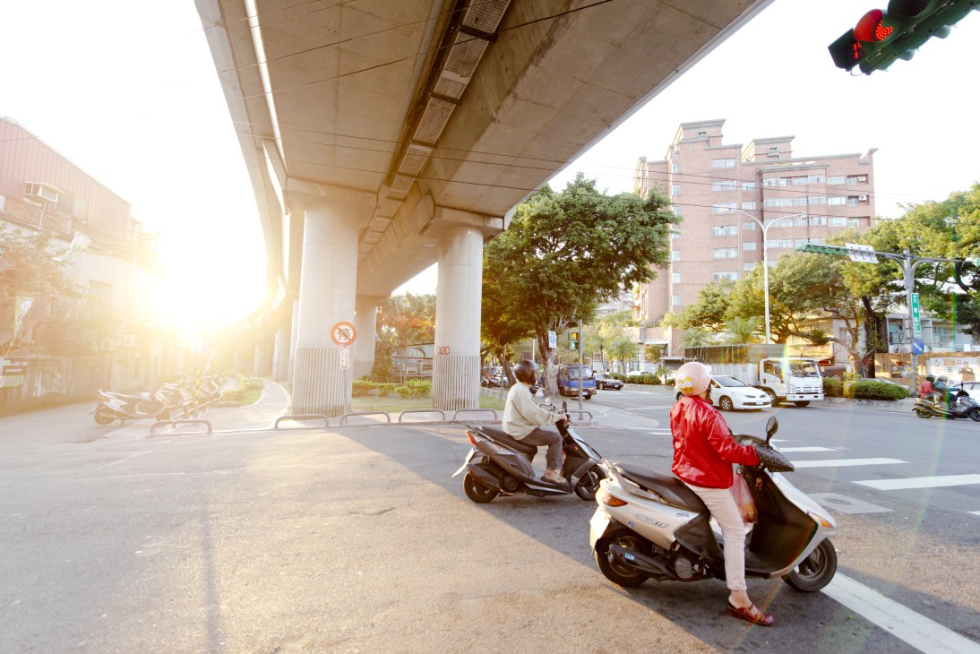 Two people on scooters waiting at a traffic light on a Beitou street under an elevated highway overpass.