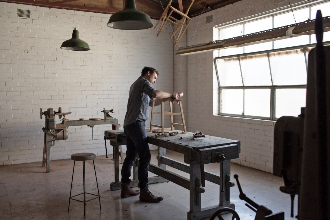Designer Benjamin Baldwin works on a wooden stool in his Melbourne workshop filled with vintage machinery and hand tools.