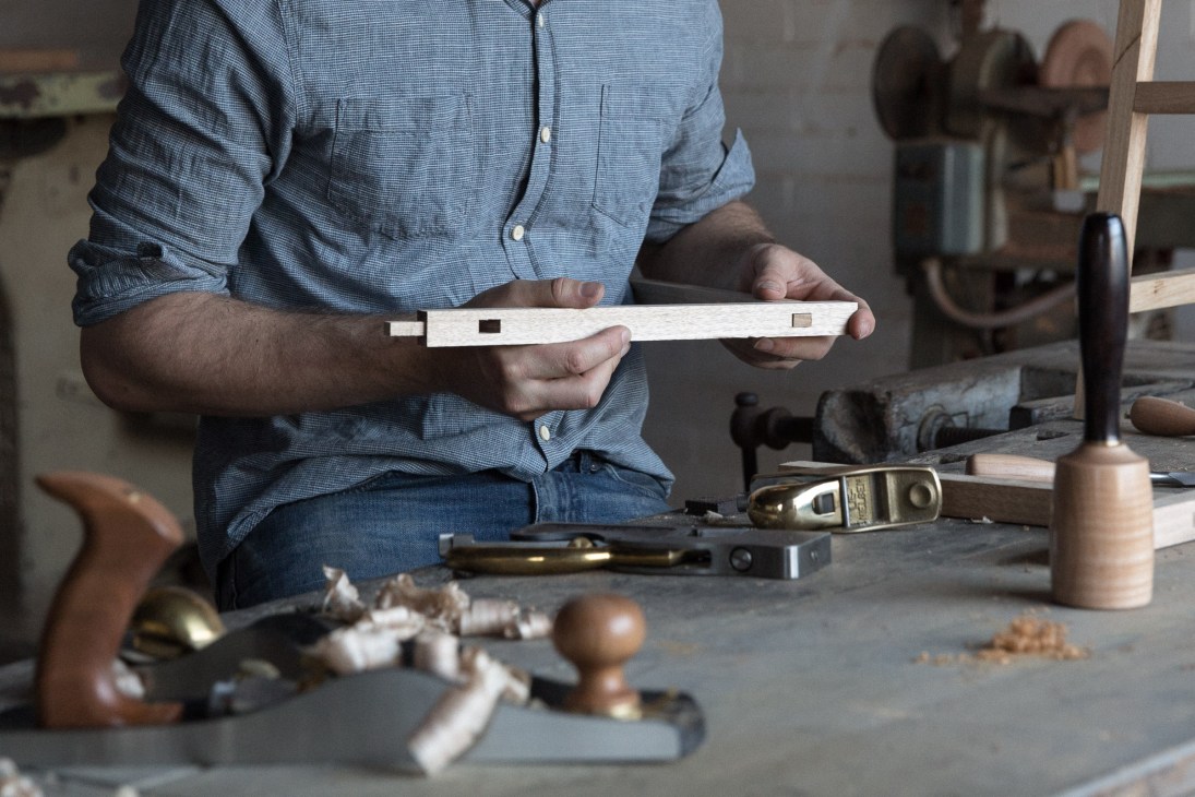 Designer Benjamin Baldwin holding a wooden joint in his Melbourne workshop surrounded by hand tools.