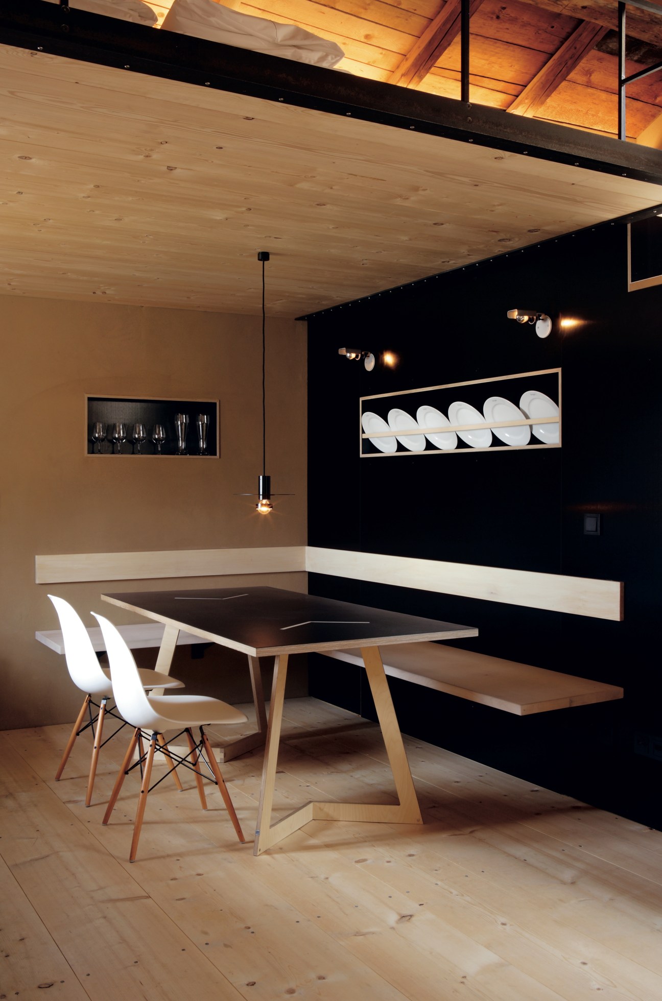 Modern dining room with wooden flooring, light wood ceiling, black accent wall displaying white plates, wooden table, white chairs, and pendant lighting.