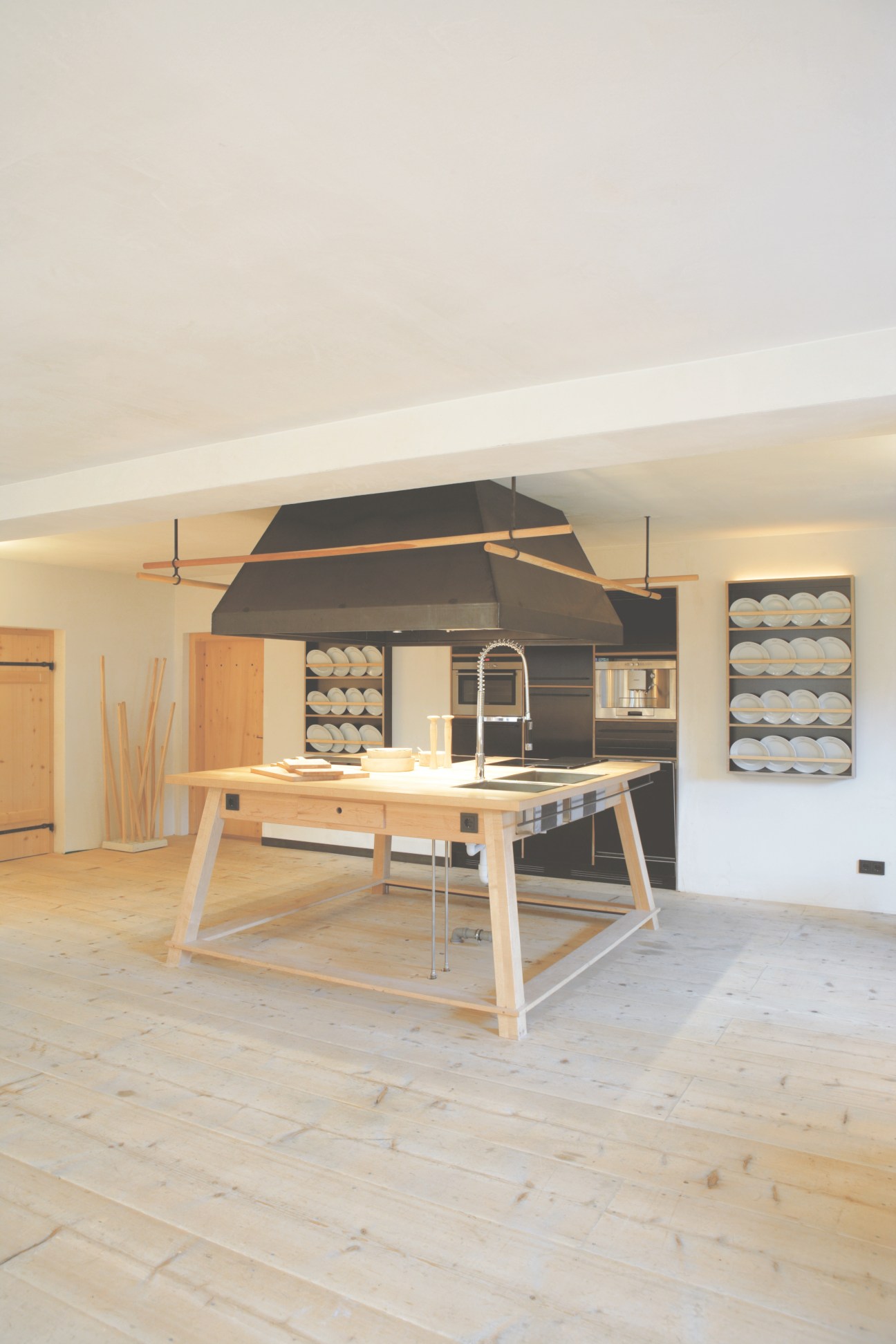 Modern kitchen interior with light wood flooring, central island, black hood vent, and open shelving displaying white plates.