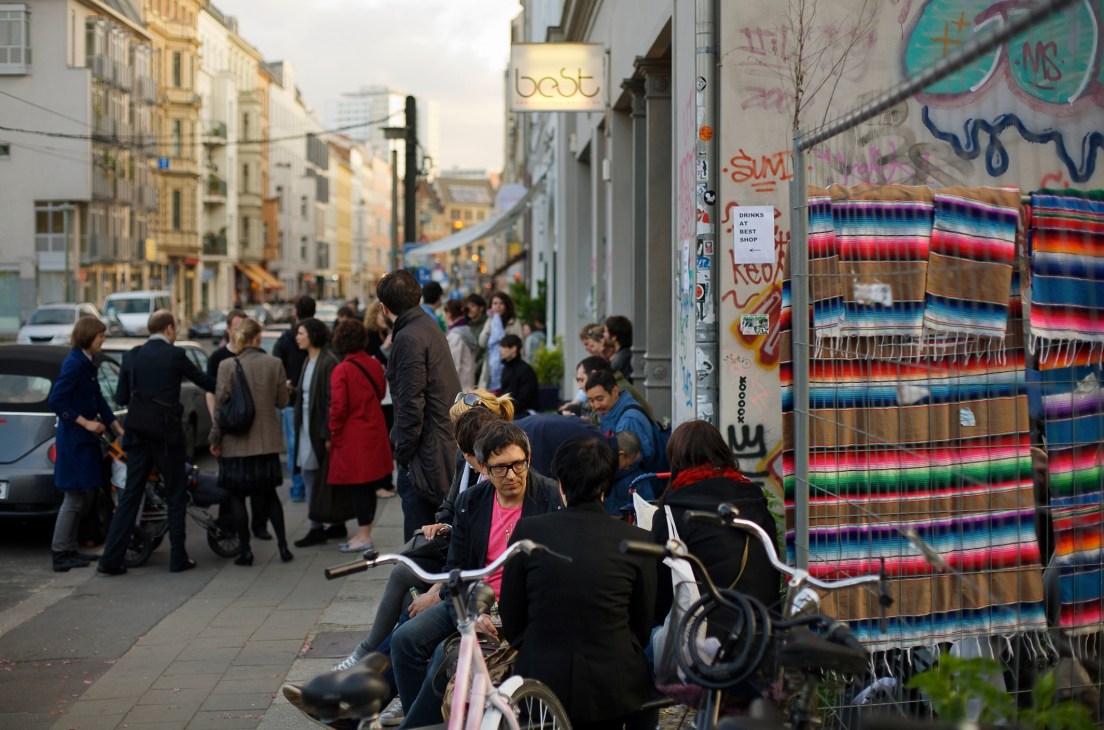Street scene on Alte Schönhauser Strasse in Berlin's Mitte district with pedestrians, graffiti-covered walls, and colorful textiles displayed for sale.