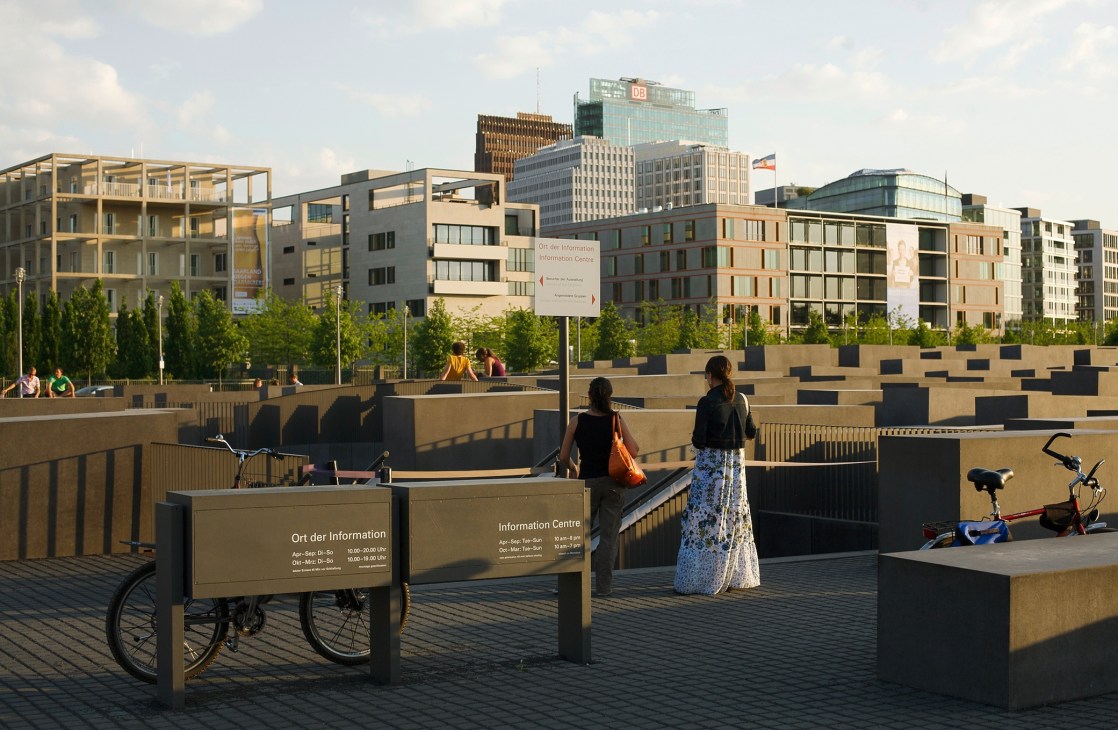 People walking on a modern plaza with geometric concrete blocks, modern residential buildings visible behind