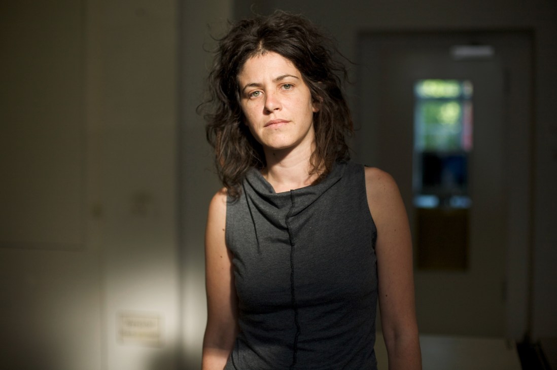 Young woman with dark shoulder-length hair wearing a dark sleeveless top in a minimalist interior space.