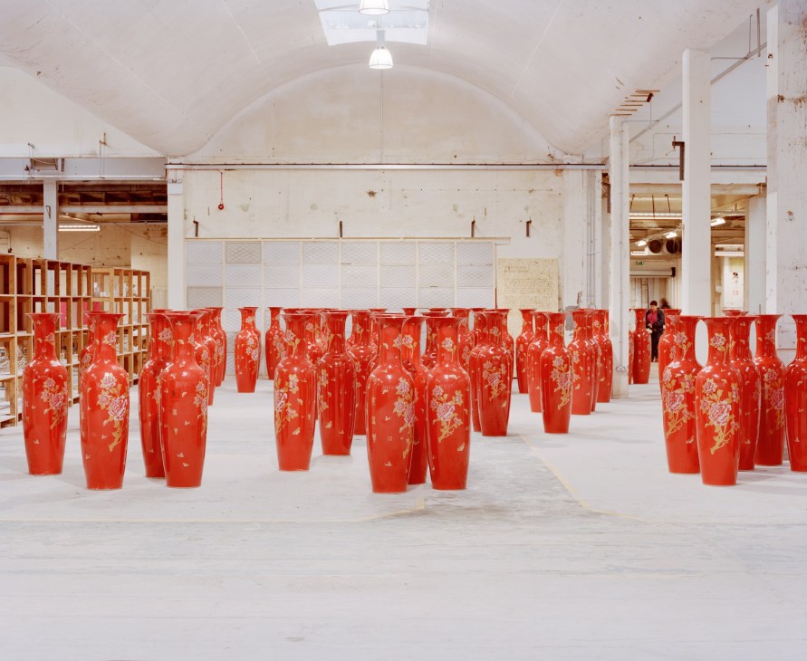 Red ceramic vases with floral designs displayed in rows inside a white industrial building with arched ceiling.