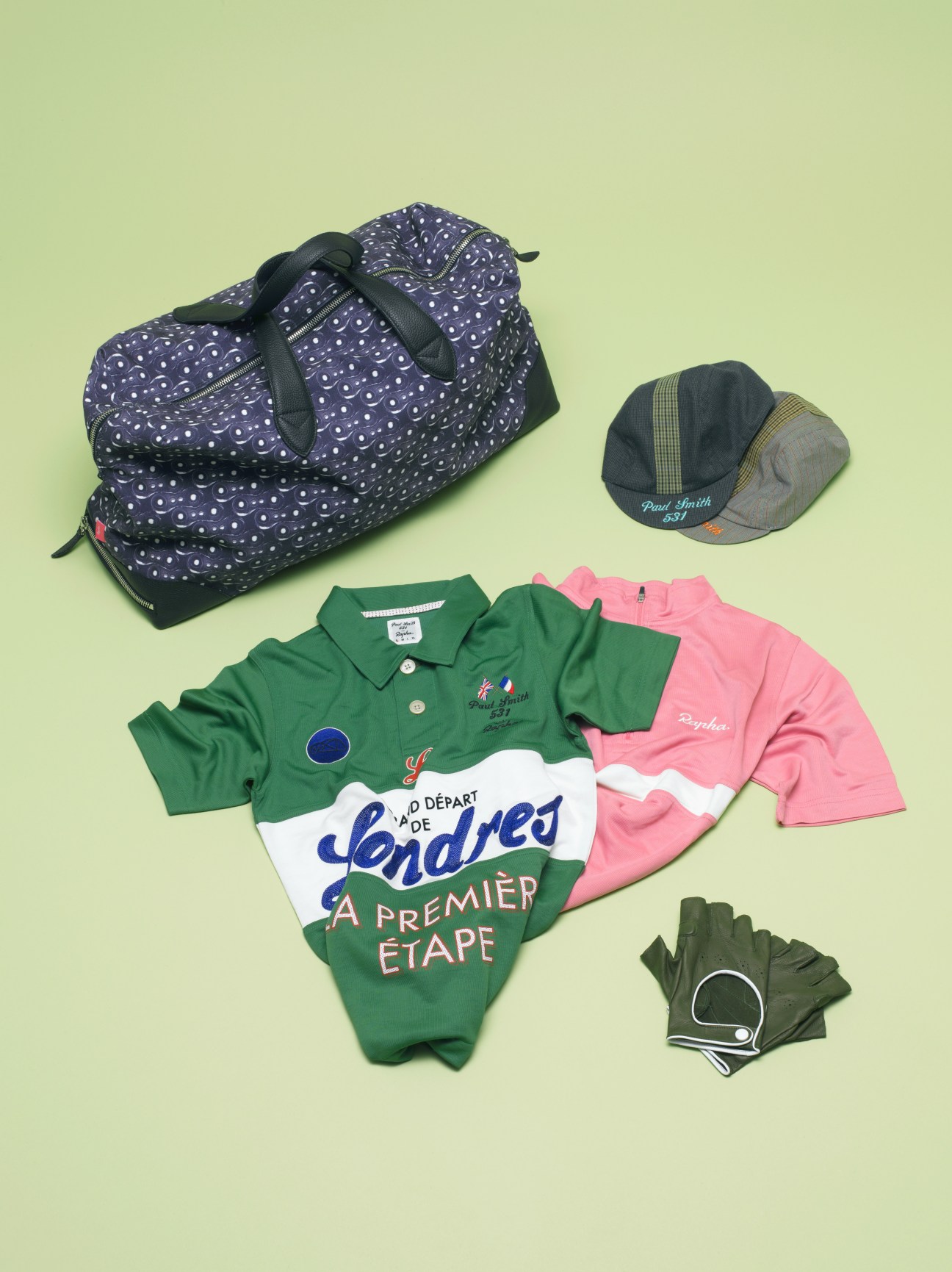 Rapha cycling apparel including jerseys, gloves, cap and patterned bag on green background.