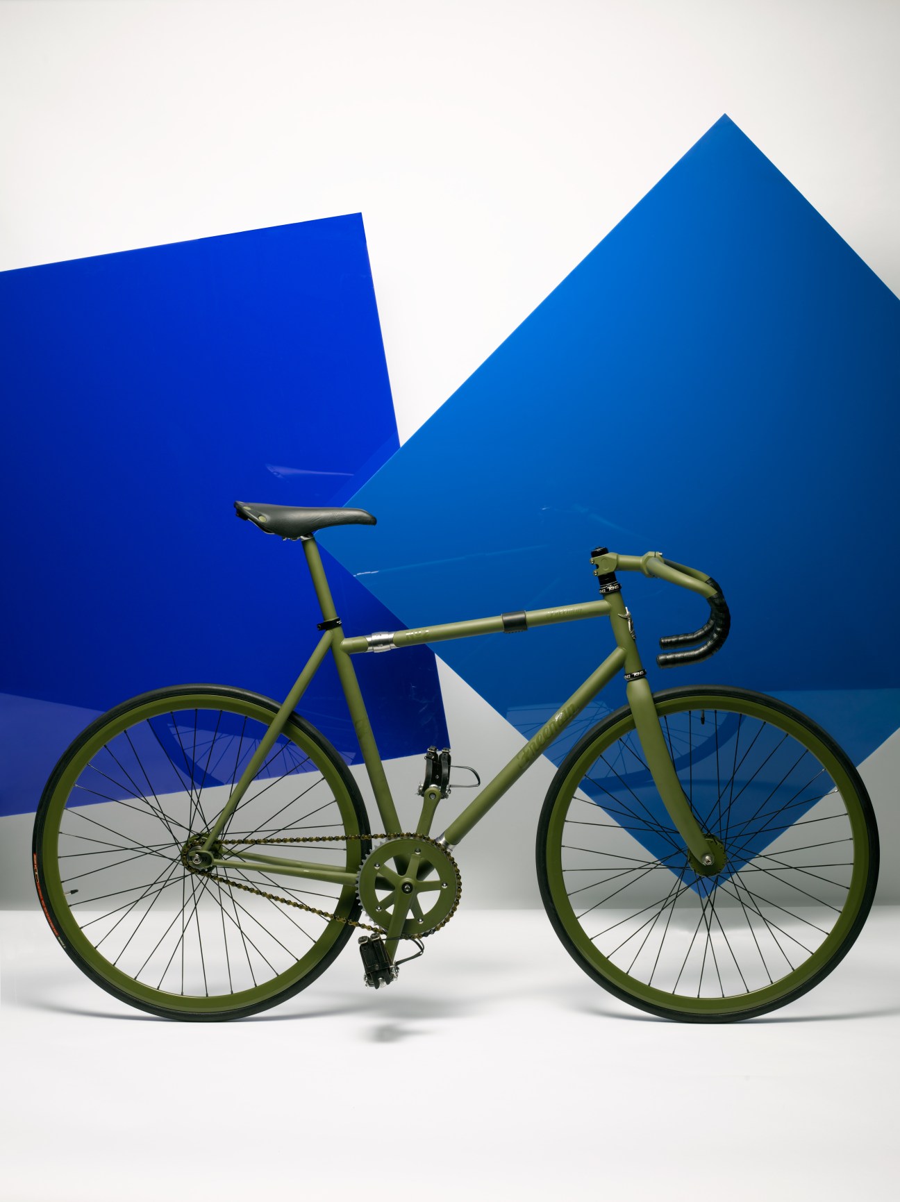 Olive-green fixed-gear track bicycle with drop handlebars against geometric blue and white background.
