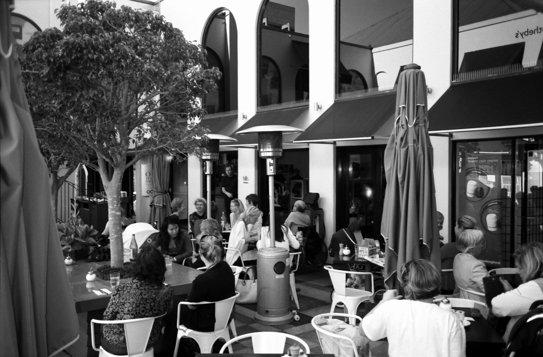 Black and white photograph of a crowded outdoor courtyard café with diners seated at tables under umbrellas and awnings.
