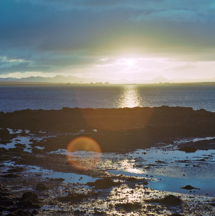 Sunset view over rocky Icelandic coastline with mountains visible across the water and golden light reflecting on the sea.
