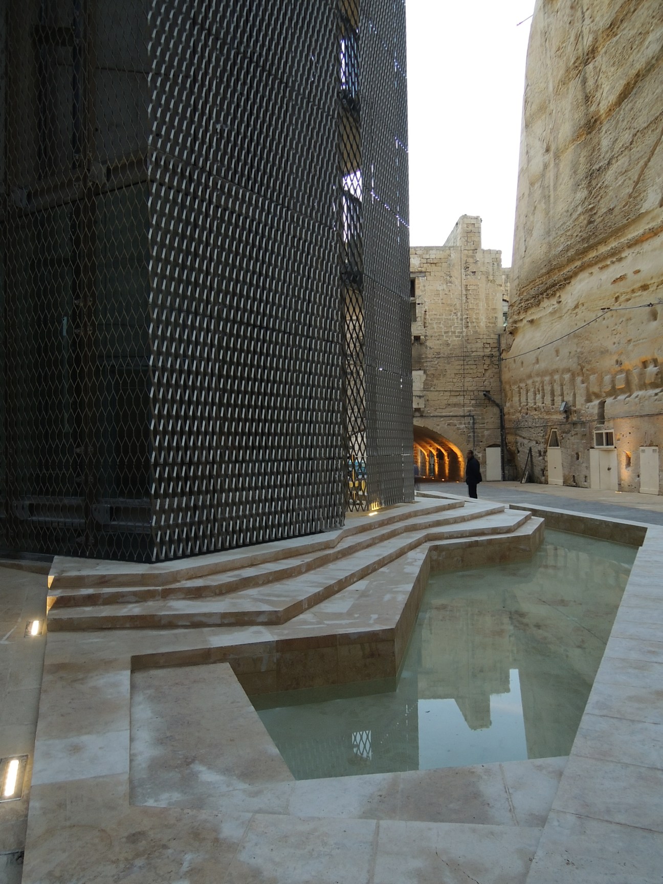 Modern lift shaft with galvanised steel mesh connecting Valletta's upper and lower town, flanked by 16th-century stone ramparts.