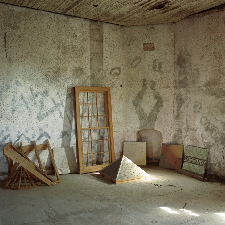 An abandoned room with deteriorating textured walls, wooden architectural fragments, a gridded window frame, and construction materials scattered on a concrete floor.