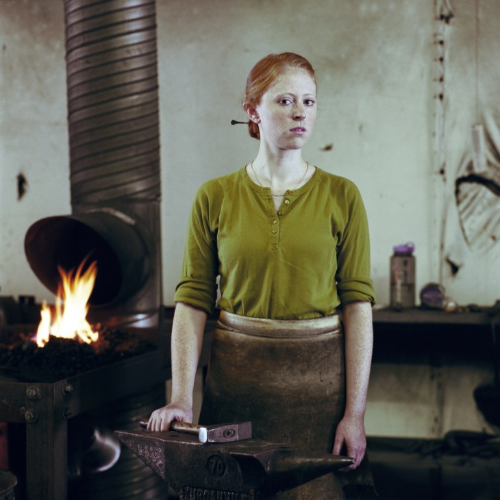 Aislinn Lewis, a student in forged architectural ironwork, stands at an anvil with a hammer in a workshop with an active forge fire behind her.