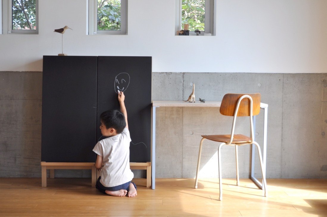 Child drawing on a black kokuban locker cupboard with a wooden desk and chair nearby in a bright room.