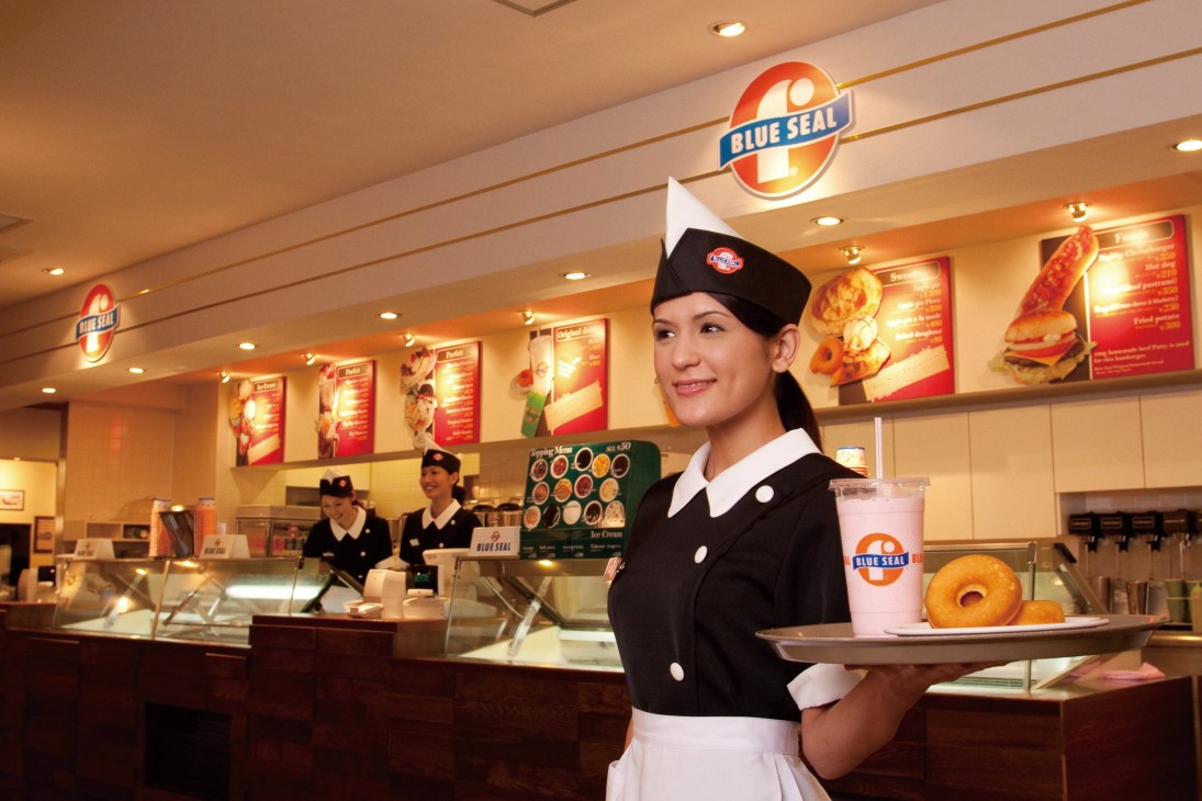 Blue Seal ice cream shop employee in uniform holding a drink and donut in a 1960s-style parlor.
