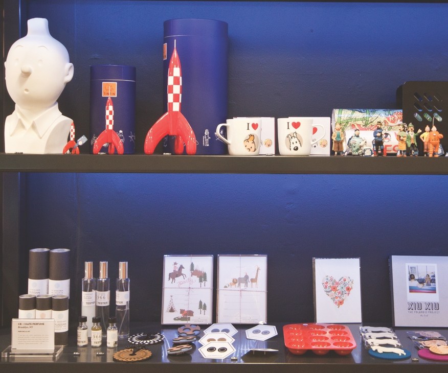 B&M store display shelves featuring design products including sculptures, red rocket toys, mugs, perfume bottles, and illustrated prints against a blue wall.