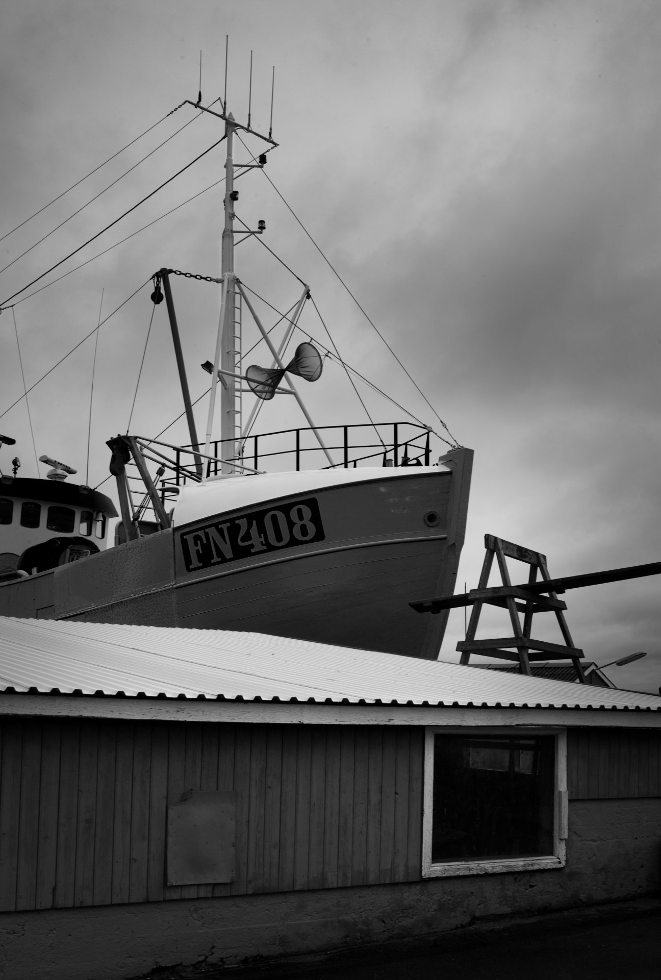 Black and white photograph of a fishing boat with registration number FN2408 docked beside a corrugated metal building on Læsø island.
