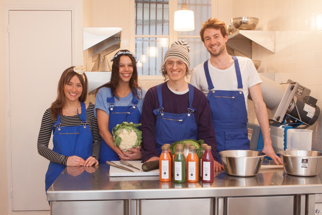 Four people wearing blue aprons stand behind a kitchen counter with fresh vegetables and bottled juices.