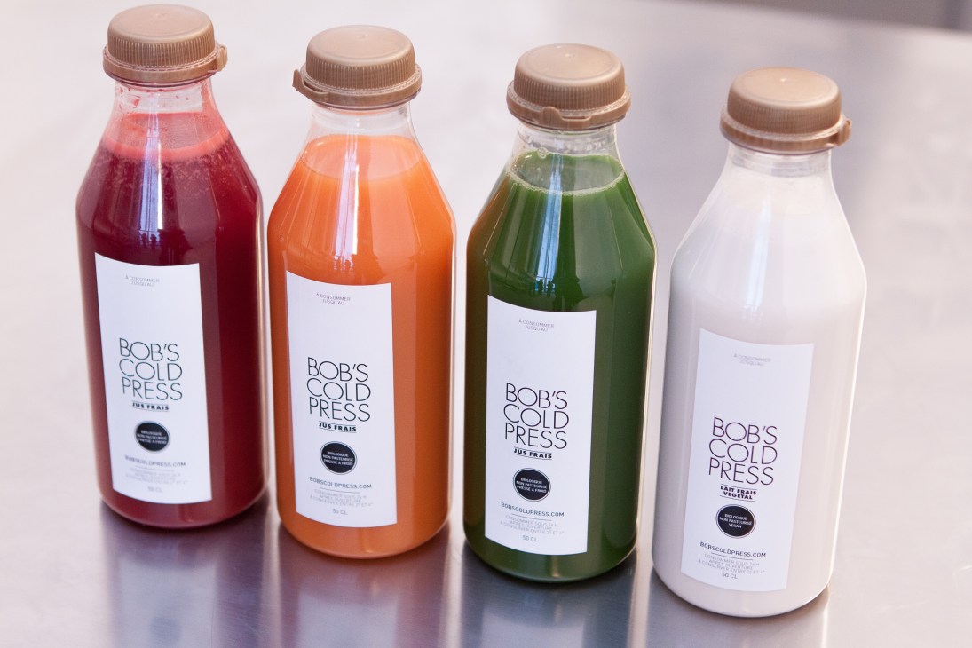 Four glass bottles of Bob's Cold Press cold-pressed juices in red, orange, green, and white varieties.