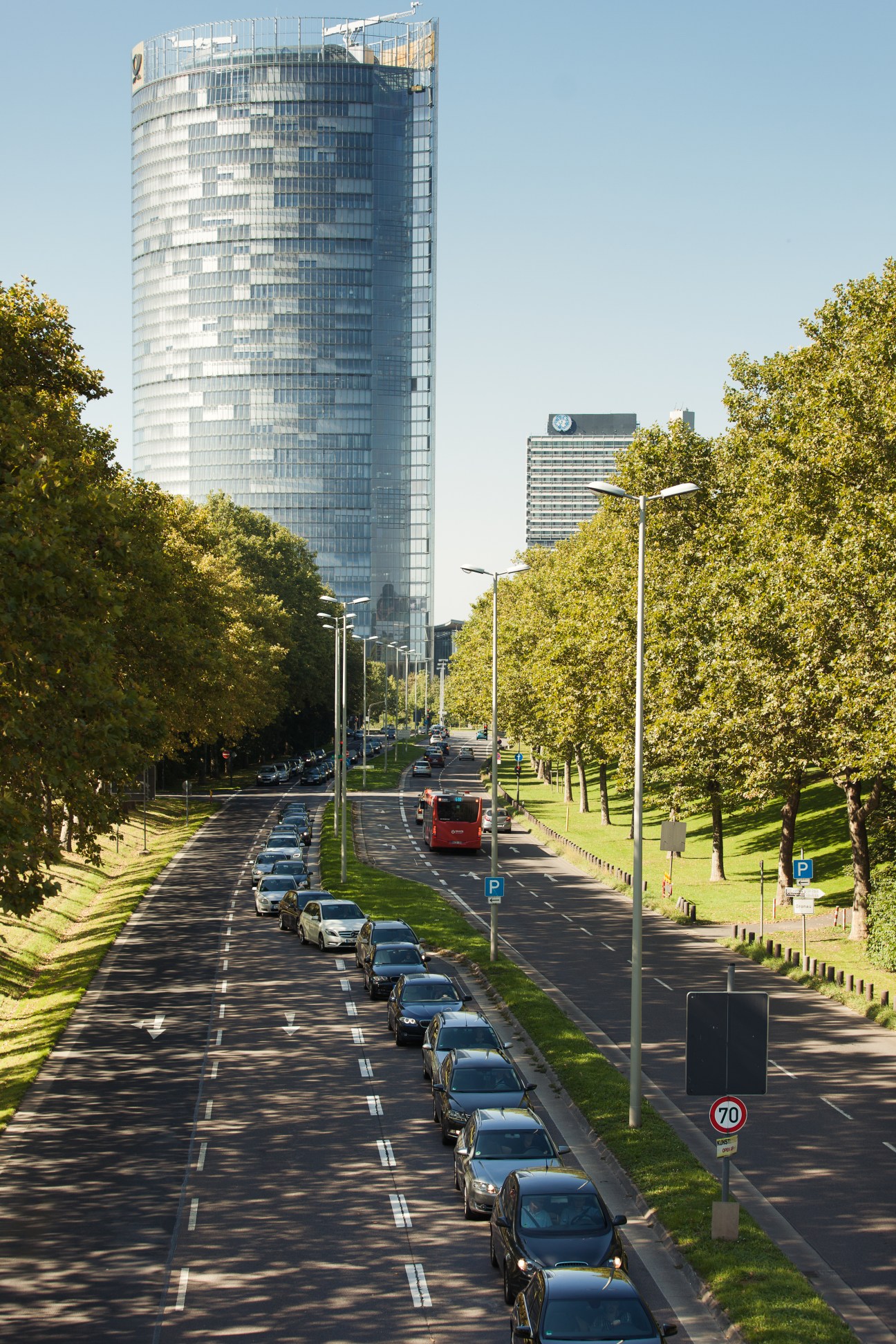 Modern cylindrical glass tower of Deutsche Post DHL headquarters with UN building behind, lined street with parked cars and trees.