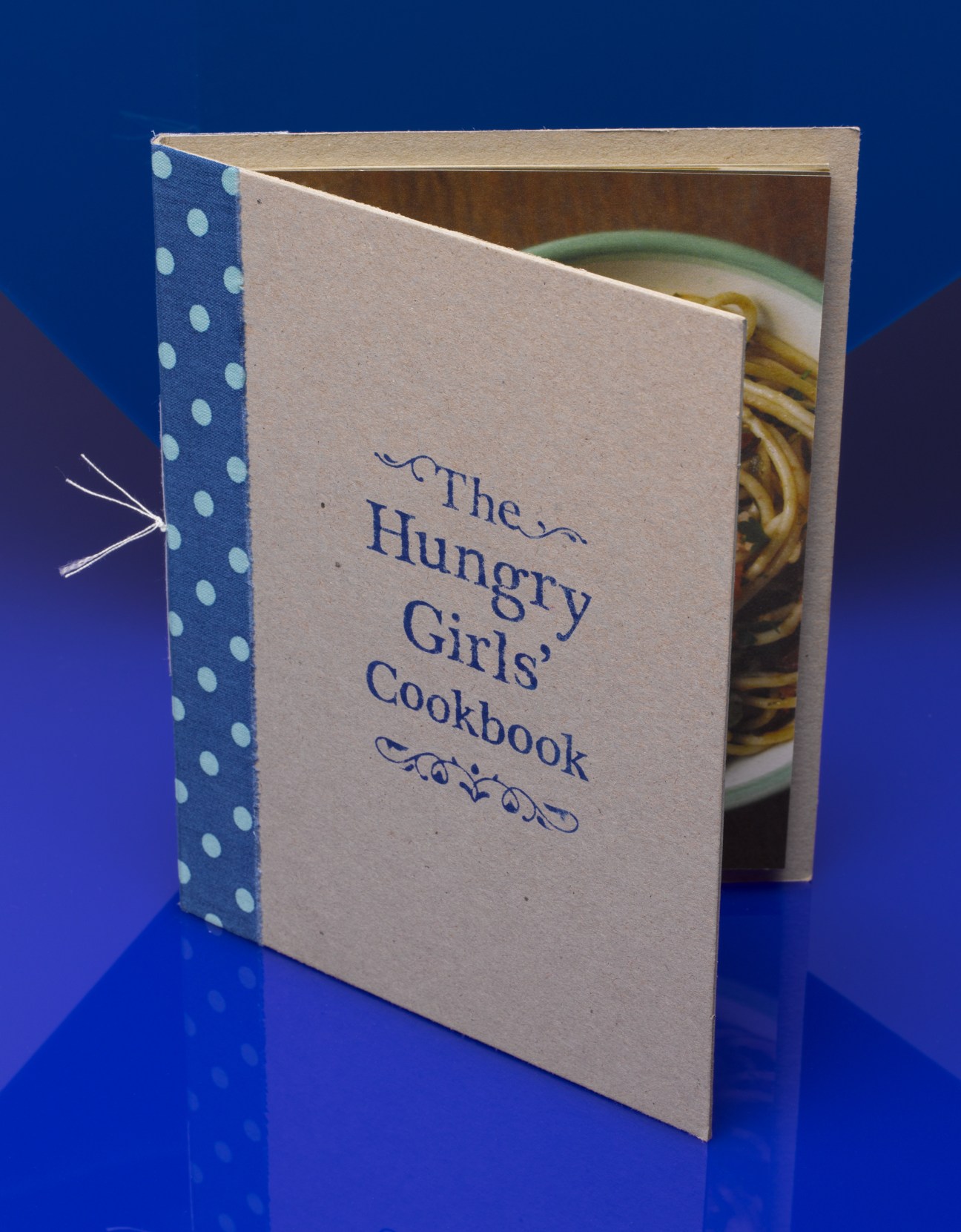 The Hungry Girls' Cookbook with hand-bound cream cover and polka dot blue spine against blue background.