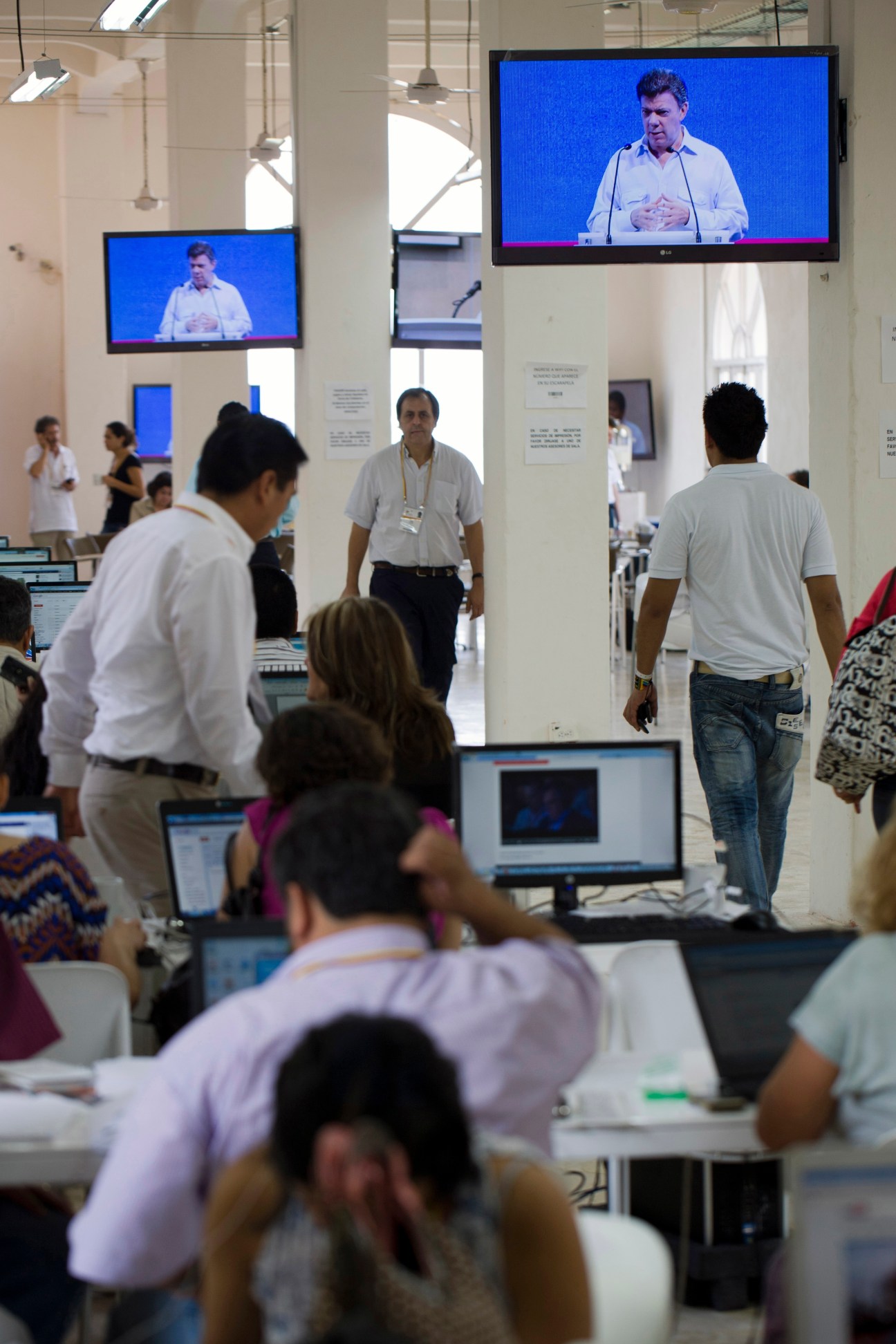 Journalists working at desks with laptops while watching a speech on overhead screens at a press center.