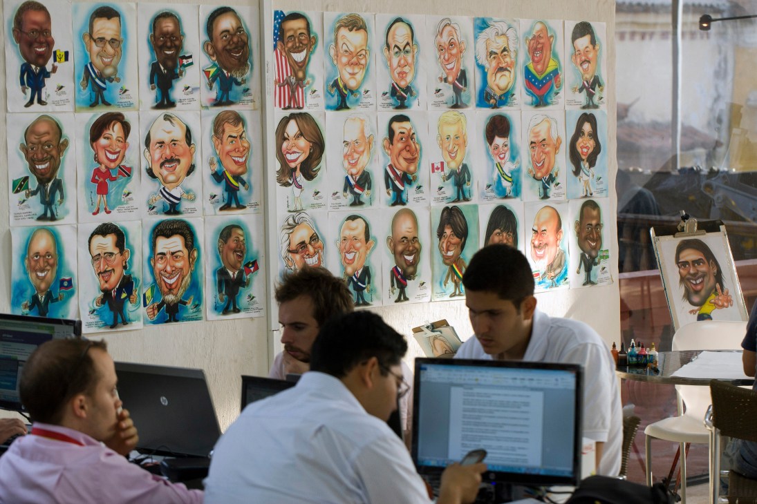 Journalists work at computers beneath a wall of colorful caricatures depicting world leaders at the Summit of the Americas press centre.