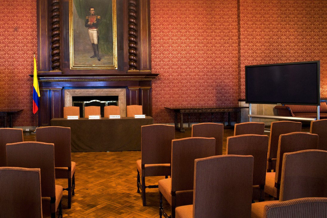 A formal conference room with orange damask wallpaper, wooden paneling, and a portrait of Simón Bolívar hanging above a fireplace.