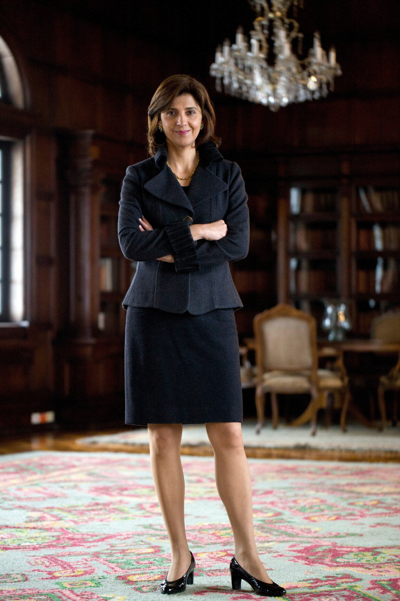Colombia's Foreign Minister María Ángela Holguín stands with arms crossed in an ornate government office with bookshelves and chandelier.